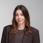 Una donna con capelli castani ondulati, che indossa un blazer marrone e collane a più strati, posa davanti a uno sfondo grigio chiaro semplice.