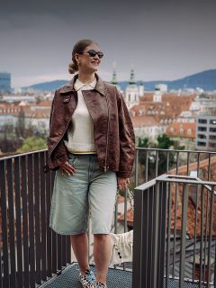 Donna con occhiali da sole e giacca marrone in piedi su un balcone con vista su un paesaggio urbano con tetti rossi e guglie di chiese in una giornata nuvolosa.