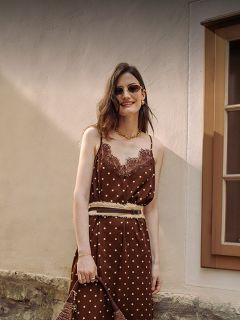Una donna in un vestito marrone a pois con bordo in pizzo, occhiali da sole e gioielli d'oro sta sorridendo davanti a una parete beige e una finestra.