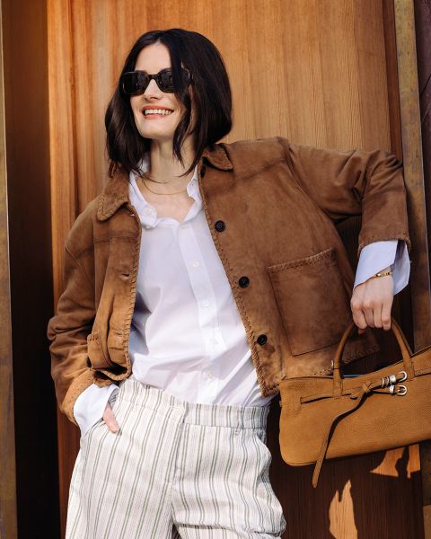 Donna con occhiali da sole che indossa una giacca marrone, una camicia bianca e pantaloni a righe, sorride e tiene una borsa marrone davanti a uno sfondo di legno.