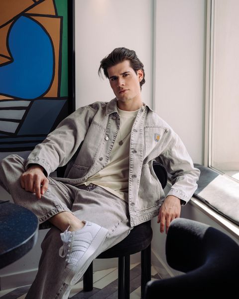 Un giovane uomo con una giacca di jeans chiara e scarpe da ginnastica bianche è seduto rilassato su una sedia vicino alla finestra in una stanza moderna con arte astratta sul muro.