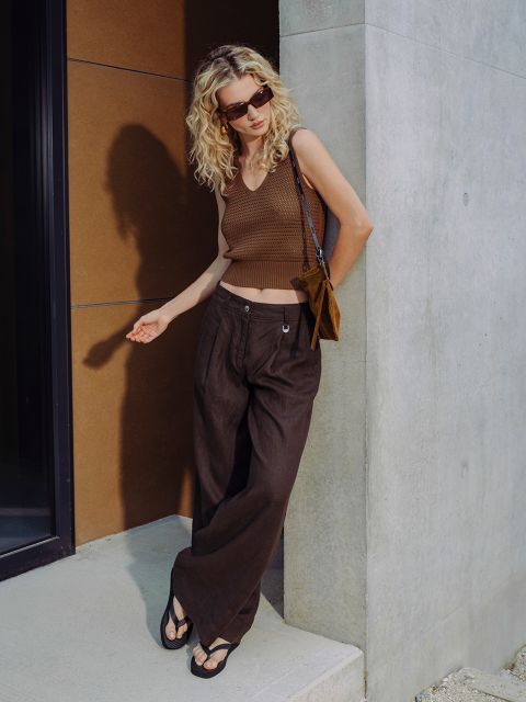 Una donna con capelli biondi ricci indossa occhiali da sole, un top marrone senza maniche, pantaloni marroni a gamba larga e sandali, e si trova di fronte a un edificio moderno.