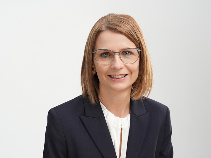 Donna con capelli lisci, castano chiaro, lunghi fino alle spalle, che indossa occhiali, un blazer blu navy e una camicetta bianca, che sorride davanti a uno sfondo chiaro e semplice.