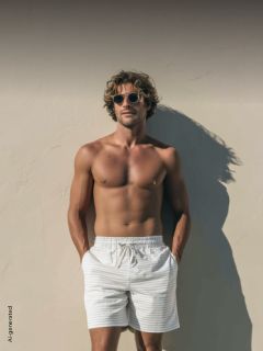 Un uomo senza maglietta, con occhiali da sole e pantaloncini a righe, sta in piedi davanti a un muro illuminato dal sole, con le mani in tasca, e proietta un'ombra.