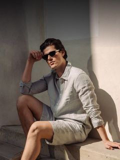 Un uomo con occhiali da sole, camicia abbottonata grigio chiaro e pantaloncini abbinati è seduto su una scalinata di cemento alla luce del sole davanti a un muro chiaro.