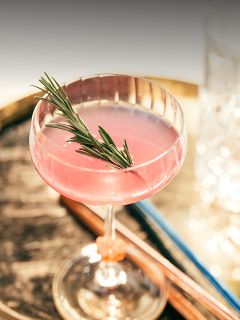 Un cocktail rosa guarnito con un rametto di rosmarino in un bicchiere coupe, su una superficie di legno rustica con bicchieri sfocati sullo sfondo.