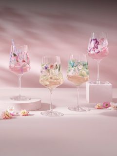 Quattro calici da vino con motivi floreali colorati, riempiti di bevande, sono posizionati su piedistalli color pastello con piccoli fiori sparsi su uno sfondo rosa.