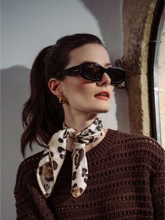 Donna con capelli scuri raccolti in una coda di cavallo, che indossa occhiali da sole neri, orecchini d'oro, un maglione marrone e una sciarpa leopardata, che guarda di lato.