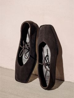 Un paio di scarpe stringate in pelle scamosciata nera, quadrate, in piedi davanti a una parete chiara e a un pavimento chiaro.