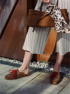 Persona che indossa pantaloni a gamba larga a righe e mocassini marroni con lacci rossi, con in mano una borsa marrone al cui manico è legato un foulard con stampa leopardata.