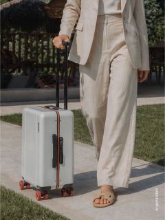 Una persona in un abito beige esce trascinando una valigia rigida bianca con ruote rosse e manico nero lungo un sentiero lastricato.