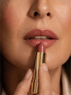 Primo piano di una donna che applica un rossetto viola sulle labbra con un tubetto di rossetto dorato.