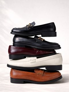 Una pila di quattro pantofole in pelle nei colori nero, bordeaux, bianco e marrone, disposte verticalmente su uno sfondo chiaro.