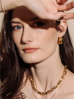 Una donna con la pelle chiara e i capelli castani, che indossa gioielli d'oro, posa con la mano che protegge parzialmente il viso dalla luce.