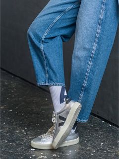 Persona in jeans blu, calzini bianchi con motivo nero e scarpe da ginnastica Veja argentate, in piedi su un pavimento maculato scuro con una gamba angolata.