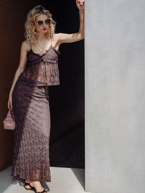 Una donna con capelli biondi ricci, che indossa occhiali da sole e un vestito di pizzo marrone, è appoggiata a un muro sotto la luce del sole splendente, con una piccola borsetta in mano.
