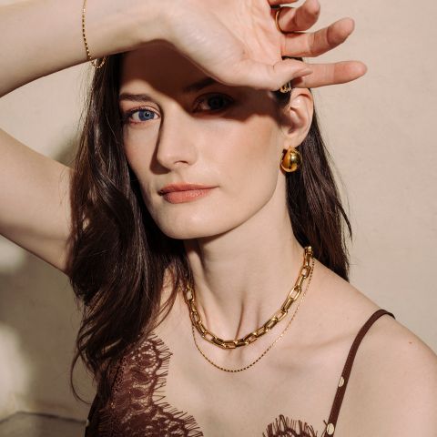 Donna dalla pelle chiara che indossa gioielli d'oro e un top di pizzo marrone, in posa con un braccio alzato per proteggere parzialmente il viso dalla luce.