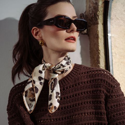 Donna con occhiali da sole neri, orecchini d'oro, un top a maglia marrone e una sciarpa fantasia legata al collo, in piedi sotto la luce del sole.