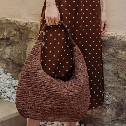 Una persona in un vestito marrone a pois tiene una grande borsa a mano marrone intrecciata, mentre sta accanto a un muro di pietra e ghiaia.