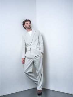 Un uomo con i capelli ricci, che indossa un abito grigio chiaro e una maglietta bianca, è appoggiato a una parete bianca in un ambiente da studio minimalista.