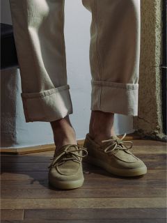 Persona con pantaloni beige con risvolto e scarpe da barca in pelle scamosciata marrone chiaro in piedi su un pavimento di legno vicino alla luce del sole.