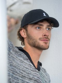 Junger Mann mit lockigem Haar und Bart, der eine schwarze Baseballmütze und einen grauen Pullover trägt und vor einem weißen Hintergrund nachdenklich zur Seite schaut.