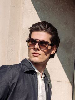 Uomo con capelli castani acconciati indossa grandi occhiali da sole quadrati, una giacca scura e una camicia chiara, e sta in piedi alla luce del sole accanto a un muro strutturato.