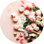 Rosa Rosen, grüne Blätter und verschiedene Macarons sind auf einer hellrosa Fläche arrangiert, mit verstreuten Blütenblättern und einem Paar Gläser in der Nähe.