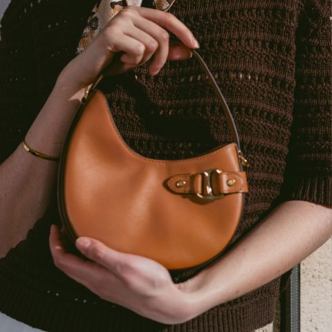 Persona con una piccola borsa a mano in pelle marrone chiaro con chiusura a fibbia, un top in maglia marrone e un braccialetto dorato.