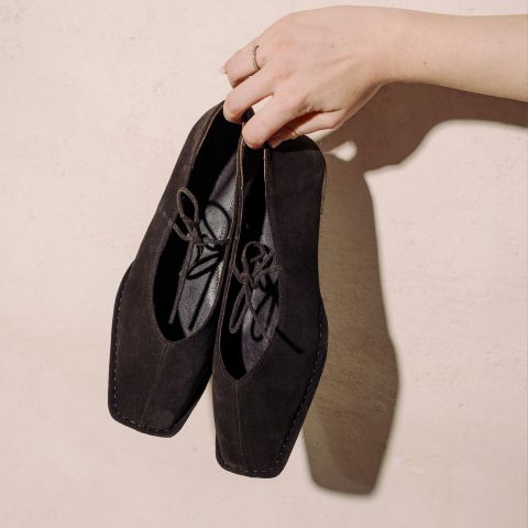 Una mano tiene un paio di scarpe stringate in pelle scamosciata nera con punta quadrata davanti a una parete strutturata beige chiaro.