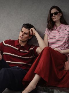 Un uomo e una donna sono seduti contro un muro, entrambi indossano polo a righe; l'uomo indossa pantaloni scuri, la donna una gonna rossa e occhiali da sole.