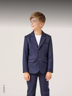 Un ragazzo in abito blu navy e camicia bianca è in piedi, sorridente e guarda di lato su uno sfondo chiaro.