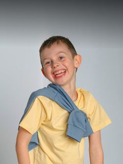 Ragazzo sorridente con una camicia gialla e un maglione blu sulle spalle, in piedi davanti a uno sfondo grigio semplice.