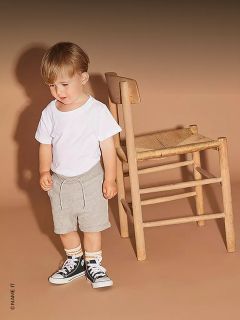 Un bambino piccolo con una maglietta bianca, pantaloncini grigi e scarpe da ginnastica nere è in piedi accanto a una sedia di legno davanti a uno sfondo beige.