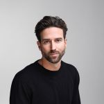 Un uomo con capelli corti castani e barba, che indossa un maglione nero, è seduto davanti a uno sfondo semplice grigio chiaro e guarda la telecamera con uno sguardo neutro.