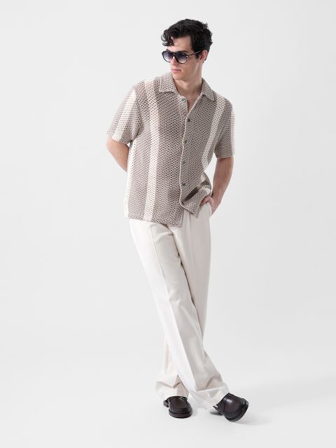 Un uomo con occhiali da sole, una camicia beige a maniche corte a fantasia e pantaloni color crema sta in piedi con una mano in tasca davanti a uno sfondo semplice.