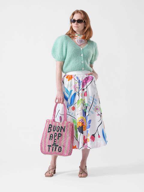 Donna con occhiali da sole che indossa un cardigan verde menta, una gonna a fiori, una sciarpa e sandali, e tiene in mano una borsa rosa con la scritta "BUON APPETITO".