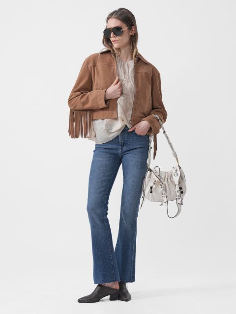 Una donna con occhiali da sole indossa una giacca marrone con frange, un top beige, jeans blu, scarpe nere e una borsa bianca, e posa davanti a uno sfondo bianco.