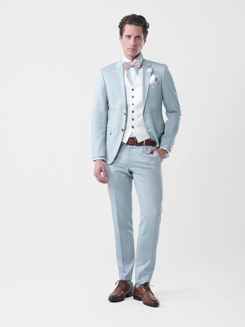 Uomo in abito azzurro, gilet bianco, papillon e scarpe marroni, in piedi davanti a un semplice sfondo bianco.