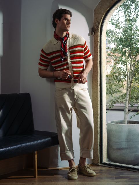 Un uomo con una polo a righe in maglia, pantaloni color crema e occhiali da sole posa alla finestra con una pianta.