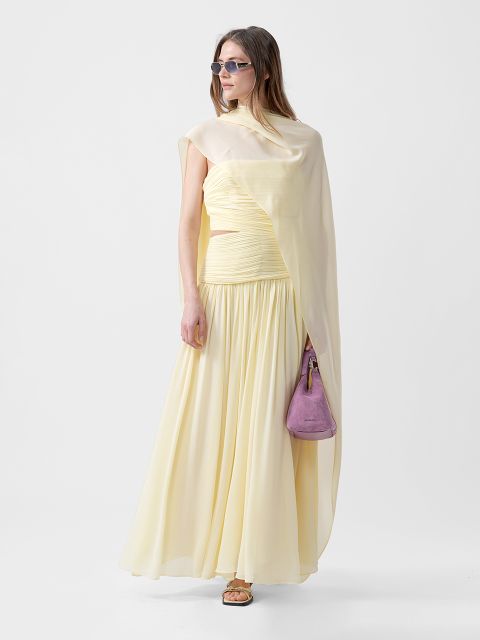 Donna in un abito giallo pallido fluente con spalla drappeggiata, occhiali da sole, sandali e una piccola borsetta viola davanti a uno sfondo semplice.