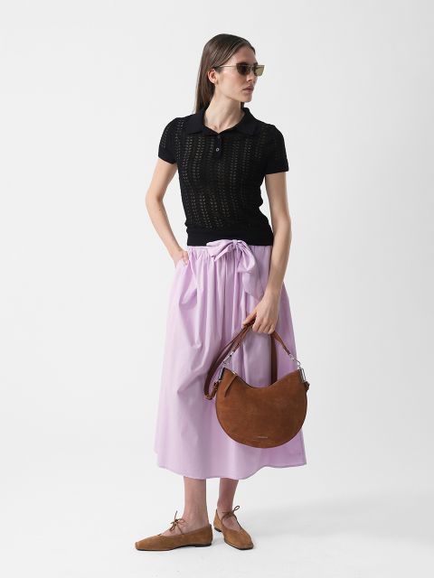 Donna con occhiali da sole che indossa un top nero a maniche corte, una gonna viola chiaro e scarpe marroni, mentre tiene una borsa a mezzaluna marrone davanti a uno sfondo bianco.