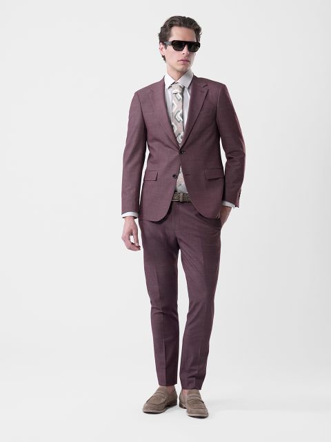 Uomo in abito viola con cravatta fantasia, camicia bianca, occhiali da sole e scarpe basse beige su uno sfondo bianco semplice.