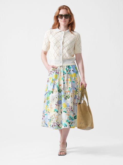 Donna con occhiali da sole, cardigan a maniche corte color crema, gonna floreale colorata, sandali e una tote bag marrone chiaro su sfondo bianco.