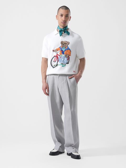 Un uomo è in piedi su uno sfondo bianco, indossa pantaloni grigio chiaro, una camicia bianca con una grafica di un orso, scarpe bicolore e una sciarpa blu-verde legata al collo.