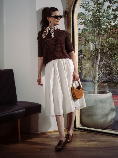 Una donna con occhiali da sole, maglione marrone e gonna bianca posa vicino a una finestra con un albero.