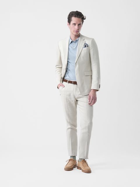 Uomo in abito crema, camicia blu, cintura marrone e scarpe marroni, in piedi davanti a uno sfondo bianco semplice mentre guarda verso la telecamera.