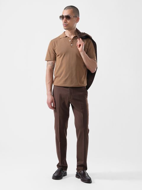 Un uomo sta in piedi davanti a uno sfondo bianco, indossa occhiali da sole, una polo marrone, pantaloni marrone scuro e tiene una giacca scura appoggiata sulla spalla.