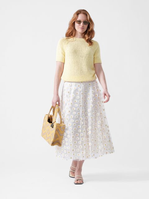 Donna con occhiali da sole che indossa un top giallo, una gonna bianca con motivo a margherite e sandali, mentre tiene una borsa in tela a fantasia gialla davanti a uno sfondo bianco.