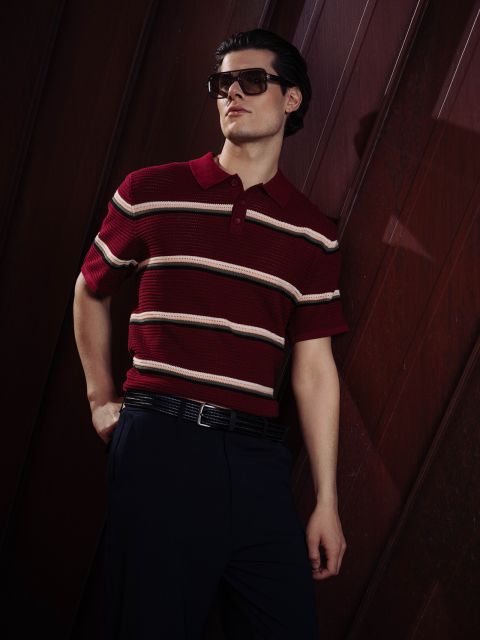 Un uomo con occhiali da sole e una polo a righe rosse posa con disinvoltura con una mano in tasca davanti a uno sfondo scuro di pannelli di legno.